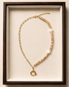 A gold chain necklace with a strand of beige and white baroque pearls, displayed in a dark wood frame with a cream velvet lining.