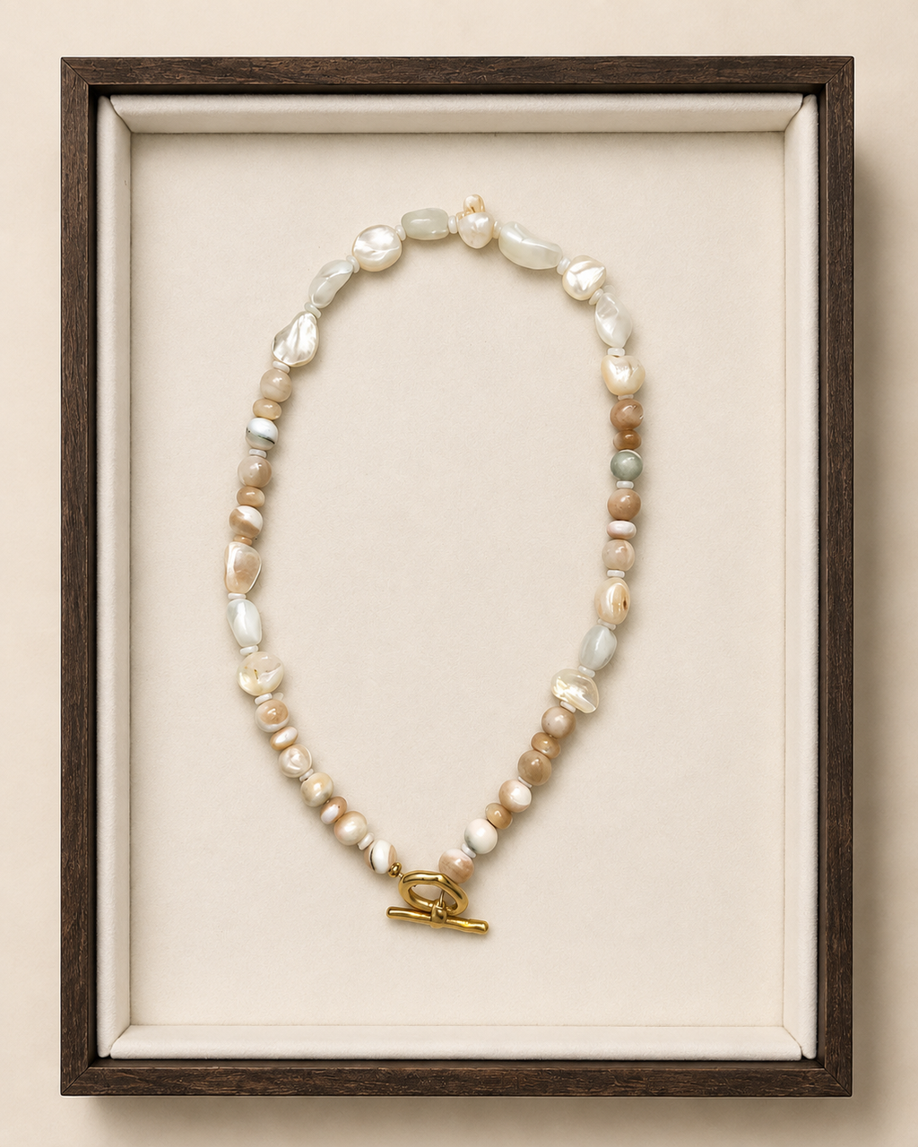 A beaded necklace with irregular white, cream, and tan beads, accented by small pale green and gray beads, and finished with a gold toggle clasp. The necklace is displayed inside a dark wood shadowbox frame with a cream velvet lining against a warm beige background.