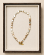 A beaded necklace with irregular white, cream, and tan beads, accented by small pale green and gray beads, and finished with a gold toggle clasp. The necklace is displayed inside a dark wood shadowbox frame with a cream velvet lining against a warm beige background.