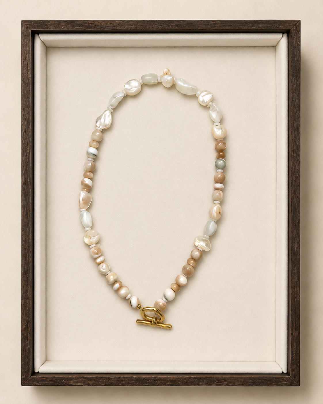 A beaded necklace with irregular white, cream, and tan beads, accented by small pale green and gray beads, and finished with a gold toggle clasp. The necklace is displayed inside a dark wood shadowbox frame with a cream velvet lining against a warm beige background.