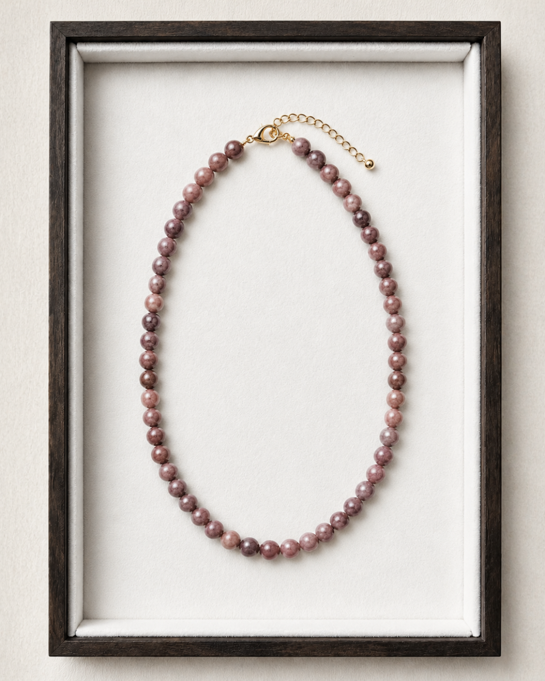 A beaded necklace with round dusty rose and purple beads, featuring a gold clasp and extender chain, displayed inside a dark wood frame with a white velvet lining.