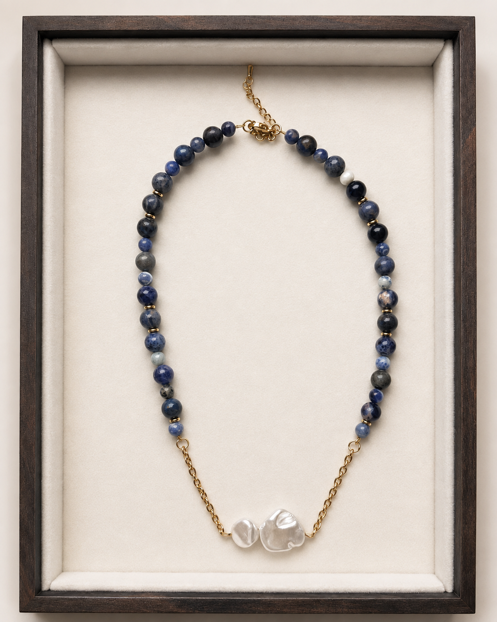 A beaded necklace with dark blue and gray beads, gold-toned chain links, and two irregular white shell-like beads at the center, displayed inside a dark wood frame with a cream velvet lining.