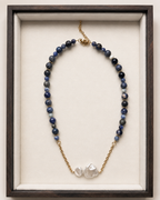 A beaded necklace with dark blue and gray beads, gold-toned chain links, and two irregular white shell-like beads at the center, displayed inside a dark wood frame with a cream velvet lining.