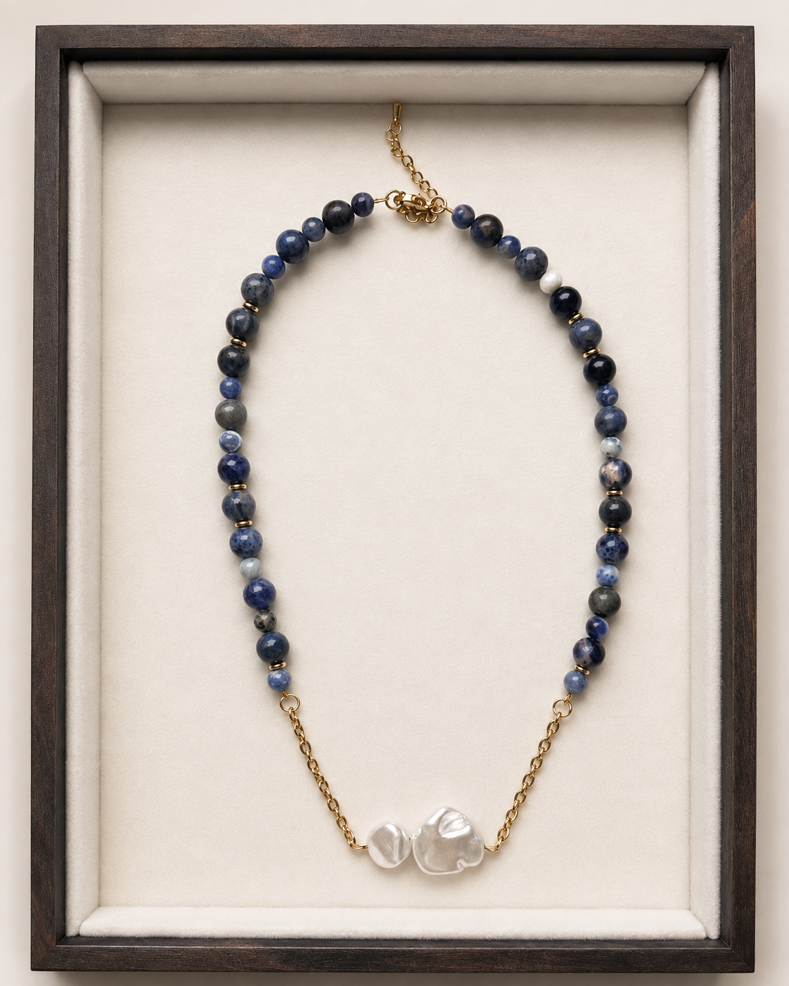 A beaded necklace with dark blue and gray beads, gold-toned chain links, and two irregular white shell-like beads at the center, displayed inside a dark wood frame with a cream velvet lining.