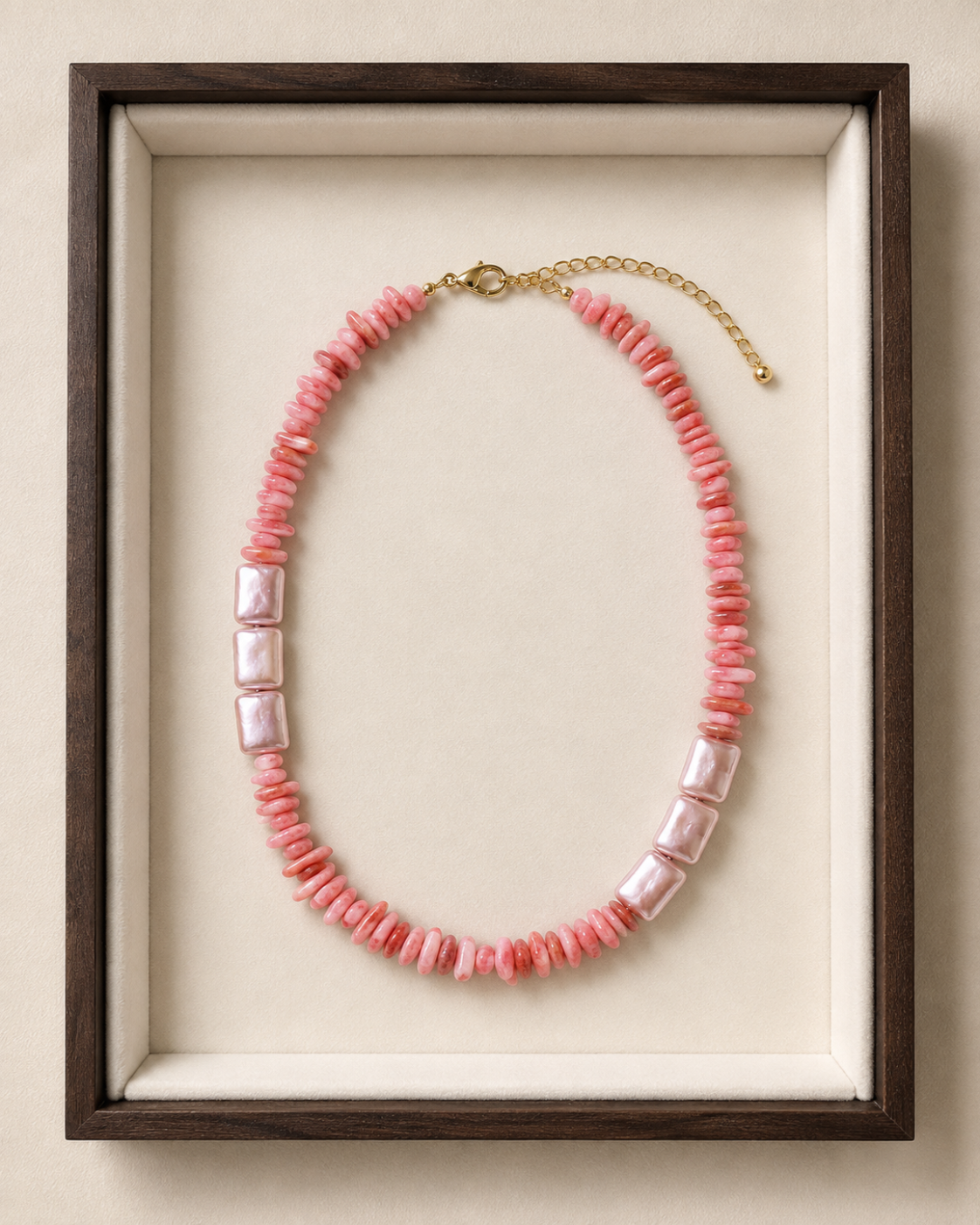A pink beaded necklace with three rectangular white pearl-like beads and a gold clasp, displayed inside a dark wood frame with a cream lining.