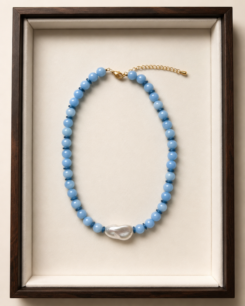 A beaded necklace with light blue round beads, a white baroque pearl pendant, and a gold chain extender, displayed inside a dark wood frame with a cream velvet lining.