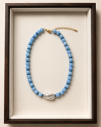 A beaded necklace with light blue round beads, a white baroque pearl pendant, and a gold chain extender, displayed inside a dark wood frame with a cream velvet lining.