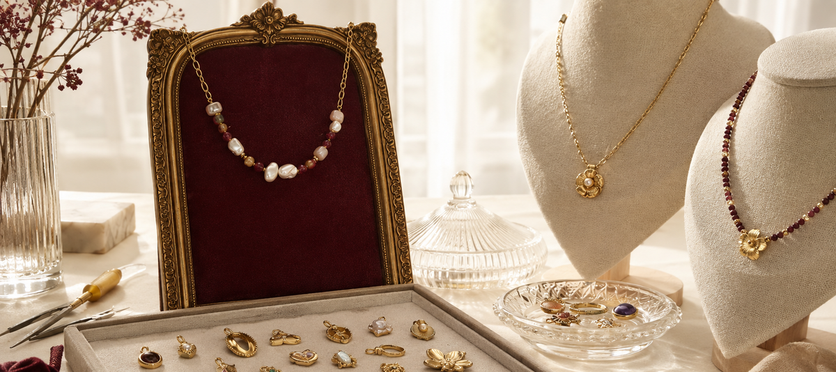 A styled jewelry display featuring multiple gold necklaces and rings on cream velvet mannequins and a gray jewelry tray, with a dark red velvet framed backdrop and soft window light in the background.